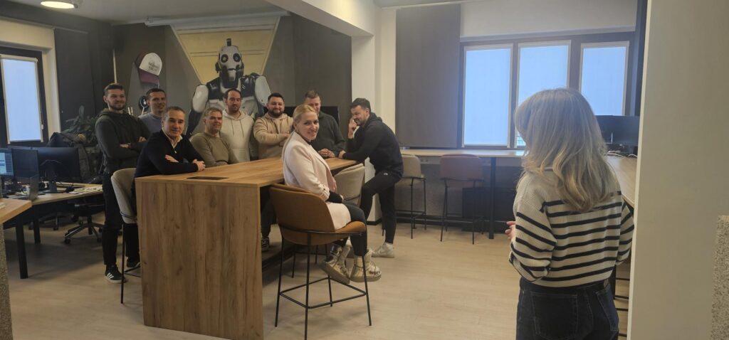 Na fotografiji se nalazi 9 sudionika edukacije o rodnoj ravnopravnosti i nediskriminaciji u sklopu projekta Re:Route. i predavačica. Tri osobe sjede za visokim stolom dok drugi stoje na nogama oko njih. Ispred njih se nalazi predavačica. Svi su ležerno odjeveni.